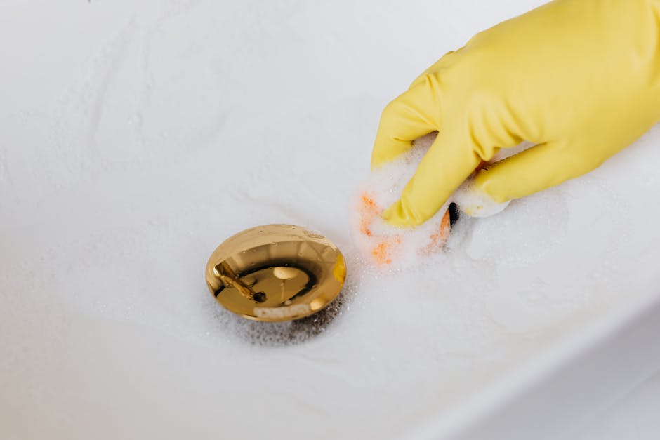 Clogged drain cleanup: a person in yellow rubber gloves using a sponge and detergent to clean a white bathroom sink from above