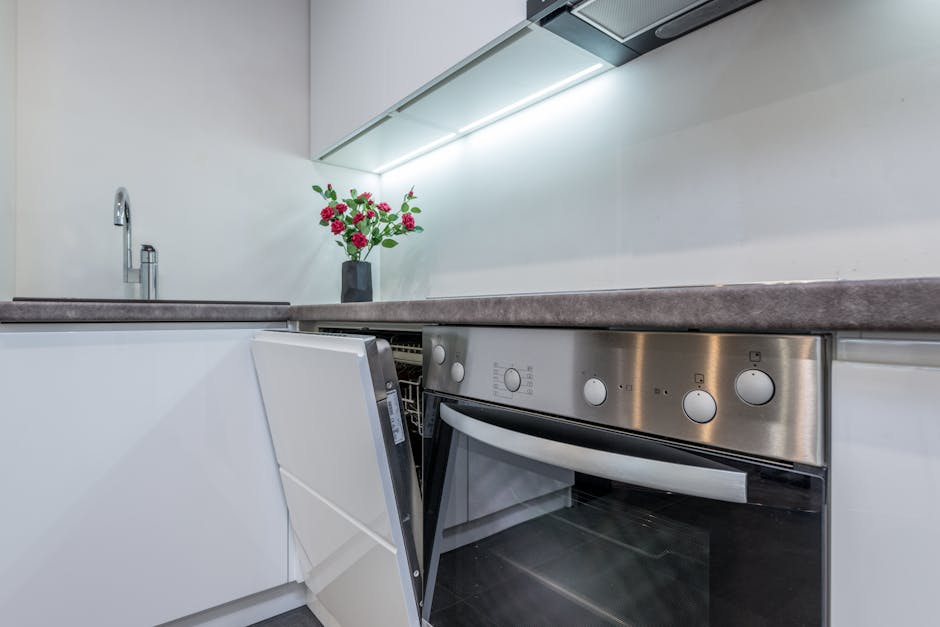 Dishwasher leaking: a standard stainless-steel residential dishwasher built into a kitchen cabinet run with a thin line of water on the tile floor at the front kickplate, the typical setup where a dishwasher leaking complaint first shows itself as a small puddle under the door